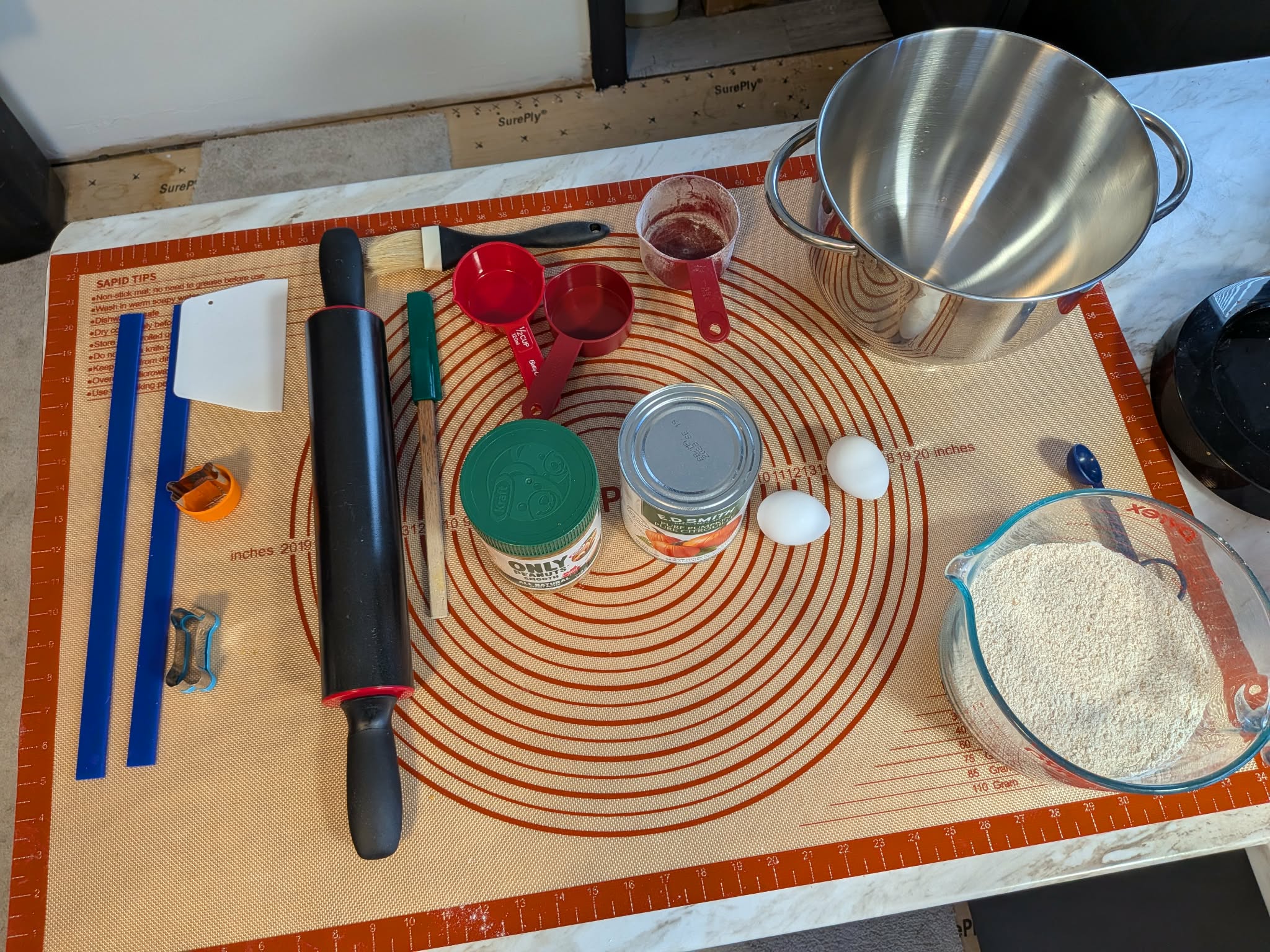 Buddy's Bones baking ingredients laid out on a silicone mat — pumpkin, peanut butter, eggs, flour and baking tools